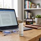 A small white bottle labeled 'Sternd' Deo Antifungal' sits on a wooden desk in a home office setting, with a laptop, eyeglasses, notebook, and plants in the background.