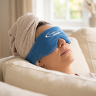 A person reclining on a beige couch with a towel wrapped around their head, wearing a blue eye mask with the brand name 'The Eye Company' printed in white.