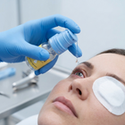 A close-up of a person receiving a drop treatment, with a gloved hand holding a small bottle and applying liquid to the eye. The person has a white patch over one eye, and the background is a clean, light gray clinical setting.
