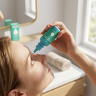 A person applying Aeon Clear contact lens drops from a teal dropper bottle to their eye in a bright bathroom setting.