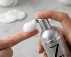 A close-up of a hand pressing the pump of a silver cosmetic bottle to dispense white foam onto another finger, with a soft blurred bathroom or studio background.