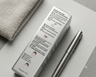 A silver metallic pen-like cosmetic tool rests beside its white rectangular box printed with black instructions and small illustrations, placed on a light gray surface next to a beige textured fabric.