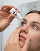 A close-up lifestyle shot of a woman applying eye drops from a white bottle labeled 'indp opt' to her eye, set against a bright, softly blurred bathroom background.