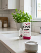 Viz Opti Preservative-Free Dry Eyes Eye Drops shown with its white and pink packaging box on a white kitchen countertop, with a blurred kitchen background featuring a potted plant.