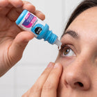A close-up of a person applying drops from a blue and white bottle labeled 'blink Immersate Plus' to their eye.
