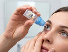 A close-up lifestyle shot of a woman applying contact lens solution from a small white and blue bottle to her eye.