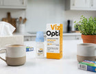 Viz Opti Preservative-Free Irritated Eye Drops shown with its orange and white box and a small dropper bottle on a kitchen counter, surrounded by mugs, tissue, and a potted plant.