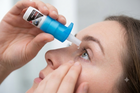 A close-up of a woman applying a blue and white eye drop from a dropper bottle to her eye, with her other hand touching her face.