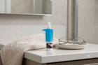 A small blue and white bottle of eye drops stands on a bathroom counter beside a folded beige towel and a pair of eyeglasses on a small dish. A mirror and tiled wall are visible in the background.
