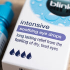 Close-up of the packaging box for Blink Intensive Soothing Eye Drops, with a blue dropper bottle partially visible on the left against a warm neutral background.