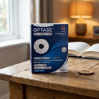 A box of OPTASE Heat Mask Pro step 1 heat masks sits on a rustic wooden table in a cozy, softly lit room with an open book and a lamp blurred in the background.