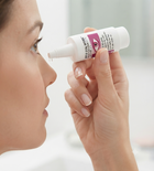 A close-up lifestyle shot of a woman holding a white and purple contact lens solution bottle near her face, with a single drop of liquid dripping from the nozzle.