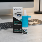 A bottle of eye drops with a bright blue cap and a matching retail box labeled 'eye doctor' and 'Screen Time Eye Drops' sit on a light gray desk in front of a blurred computer keyboard and monitor.