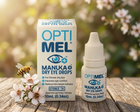 OptiMel Manuka Dry Eye Drops shown with its white and blue retail box and matching dropper bottle on a wooden surface, with a bee and white blossoms against a soft blurred outdoor background.