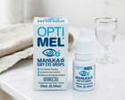 OptiMel Manuka Dry Eye Drops shown with the retail box, dropper bottle, and a glass of water on a light marble surface with a folded beige cloth in the background.