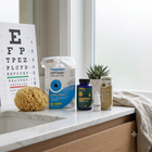 A wellness product set arranged on a white marble countertop in a bright bathroom or spa setting. The set includes an Optase eye care box, a dark blue bottle, a small white dropper bottle, and a Nicosan box, alongside a sponge, a small potted succulent, and a beige towel.