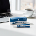 HYLO NIGHT eye drops packaging on a desk with a laptop and coffee cup in the background