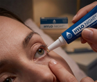 A close-up of a person applying HYLO NIGHT eye drops with a blue and white tube held by a hand, with the product box blurred in the background.